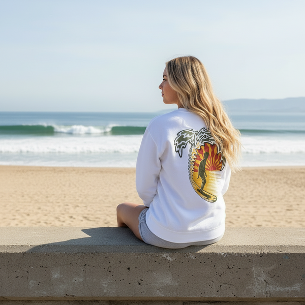 Woman sitting on a ledge at the beach wearing a white long-sleeve shirt with a colorful design.